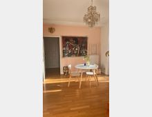 Helles Wohnzimmer mit Holzfußboden, einer rosafarbenen Wand und einer offenen Tür zu einem angrenzenden Raum. Im Vordergrund steht ein runder Tisch mit zwei Stühlen, darüber ein Kronleuchter; an der Wand hängt ein farbiges, abstraktes Gemälde.