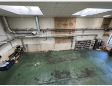 Industriehalle mit hoher Decke, sichtbaren Lüftungsrohren und einer gemischten Wand aus weißer Farbe und freiliegendem Ziegel. Auf dem abgenutzten grünen Boden stehen Regale mit Farbdosen, ein Computerarbeitsplatz und verschiedene Werkstattmaterialien.