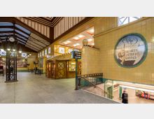 Historische U-Bahn-Station mit gelb gekachelten Wänden, einem überdachten Bereich mit verzierten Decken und einem großen Wandgemälde. Im Vordergrund ein Treppenabgang zu den Gleisen, ein alter Fahrkartenschalter aus Holz und Wegweiser zu den Bahnsteigen.