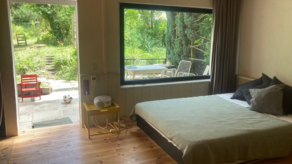 Schlafzimmer mit Holzfußboden und Holzdecke, einem Doppelbett neben einem großen Panoramafenster mit Vorhang. Links führt eine offene Tür auf eine Terrasse und in einen grünen Garten; draußen sind Sitzmöbel sichtbar.