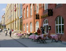 Straßenszene mit einem roten Backsteingebäude, auf dessen Fassade "12 Apostel" geschrieben steht, und mehreren Tischen mit rot-weiß karierten Tischdecken auf dem Gehweg. Im Hintergrund sind weitere Gebäude mit gläsernen Fassaden und Personen zu sehen, die die Straße entlanglaufen.