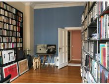 Ein Arbeitszimmer mit hohen Decken, einer blauen Wand und Holzboden. Links steht ein großes Bücherregal mit Büchern und gerahmten Bildern, während rechts ein Regal mit Aktenordnern und Büchern zu sehen ist. In der Mitte befinden sich ein kleiner Tisch mit Hockern und eine geöffnete weiße Tür, die in einen weiteren Raum führt.
