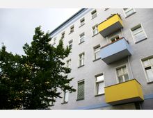Fassade eines mehrstöckigen Wohngebäudes mit hellgrauer Putzwand, weißen Fensterrahmen und zwei Balkonen in gelber und blauer Farbe. Im Vordergrund steht ein Baum mit dichtem grünen Laub.