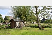 Holzfass-förmige Gartenhütte auf einer grünen Wiese mit Bäumen im Hintergrund, umgeben von einem niedrigen Holzzaun. Im Hintergrund ein Landhaus und weitere Vegetation.
