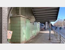 Straßenszene unter einer Hochbahntrasse mit grün gekachelten Wänden und Metallverstrebungen. Fahrräder sind an einem Geländer angeschlossen, und im Hintergrund sind Gehwege, eine Straße und Fußgänger zu sehen.