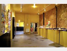 Ein Innenraum mit gelb gestrichenen Wänden und einer freiliegenden roten Backsteinwand auf der rechten Seite. Der Raum verfügt über eine lange gelbe Theke, eine Wand mit Broschürenhaltern links, sowie eine gläserne Eingangstür im hinteren Bereich.