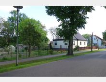 Straßenszene in einer Wohngegend mit einem kleinen, eingeschossigen weißen Haus mit Satteldach, einem Garten, der von einem Zaun umgeben ist, und vereinzelten Bäumen entlang des Gehwegs. Im Hintergrund sind weitere Wohnhäuser und dichte grüne Bäume sichtbar.