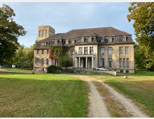 Historisches Herrenhaus mit einer verwitterten Fassade, großen Sprossenfenstern und einem steilen Mansardendach, umgeben von einem weitläufigen Rasen und Bäumen. Ein schmaler Schotterweg führt zur Eingangstreppe mit Säulenportikus.