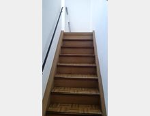 Holztreppe mit lackierten Stufen und braunem Parkettmuster, die zu einem hell beleuchteten oberen Bereich führt. Rechts ist eine weiße Wand und links ist ein schwarzes Metallgeländer zu sehen; im oberen Bereich lehnt eine Leiter an der Wand.