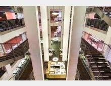 Innenansicht eines modernen, mehrstöckigen Gebäudes mit offenen Galerien, Metallgeländern und Holztreppen. Im Erdgeschoss befindet sich ein Ess- oder Cafébereich mit Tischen, Stühlen und einer zentralen Theke; hängende runde Leuchten sorgen für Beleuchtung.