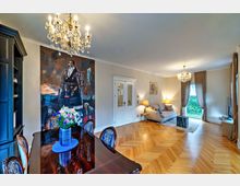 Wohnzimmer mit eleganter Einrichtung: Ein Raum mit Parkettboden, einem großen Gemälde an der Wand, einem Kristall-Kronleuchter, einem Esstisch im Vordergrund und einer gemütlichen Sitzecke mit Sofa und Vorhängen im Hintergrund. Große Fenster lassen viel Tageslicht herein.