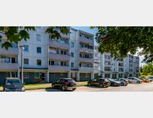 Mehrstöckiger Wohnblock aus heller Fassade mit Balkonen, davor mehrere geparkte Autos und ein Gehweg, umgeben von großen, teils belaubten Bäumen.