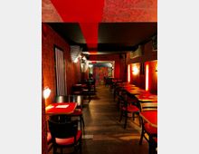 Ein Restaurant mit roten Tischen und Stühlen, umgeben von dunkelroten, verzierten Wänden und warmem Licht. Im Hintergrund ist eine bunt gemusterte Wand zu sehen, während die Decke ebenfalls detaillierte Muster und rote Streifen aufweist.