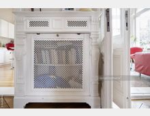 Weißes, verzierter Heizkörperverkleidung im klassischen Stil mit dekorativem Gitter, umgeben von einem hellen, modernen Innenraum mit weißen Wänden, Holzboden und roten Sitzmöbeln im Hintergrund.