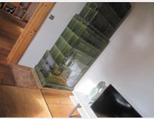 Ein Wohnraum mit einem traditionellen, grün glasierten Kachelofen an einer weißen Wand, daneben ein Fernseher auf einem kleinen Tisch. Der Boden besteht aus Holz und Fliesen, und im Hintergrund ist eine offene Tür mit einem Holzrahmen sichtbar.