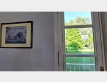 Ein Rahmenbild mit der Aufschrift „Schloss Feldow“ hängt an einer weißen Wand links im Bild, während rechts ein großes Fenster mit weißen Rahmen den Blick auf eine grüne Wiese und Bäume im Sonnenlicht freigibt.