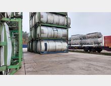 Ein Lagerplatz mit gestapelten Tankcontainern aus Metall in grünen Rahmen, daneben ein Zugwaggon mit weiteren Tankcontainern. Im Hintergrund sind Bäume und vereinzelt Container zu sehen.