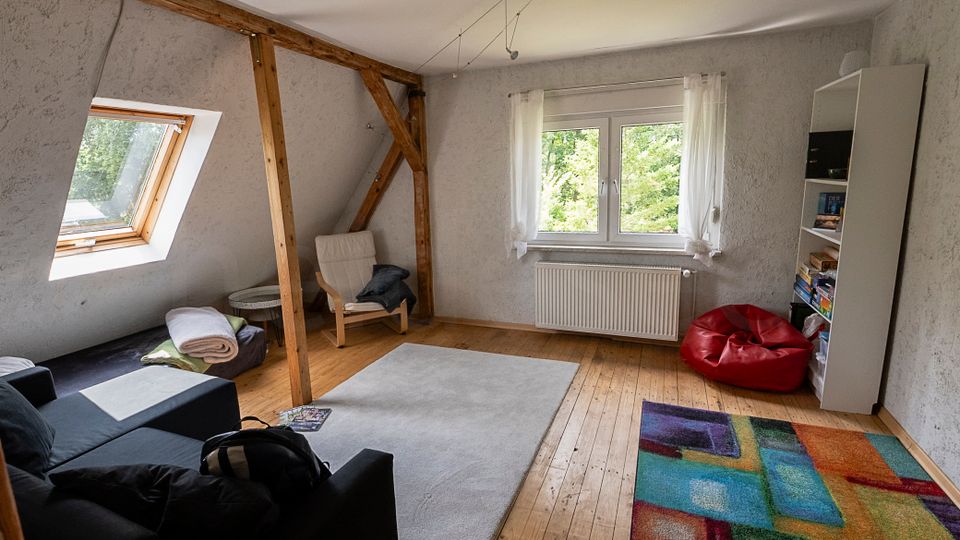Helles Dachzimmer mit schrägen Wänden, sichtbaren Holzbalken und Holzfußboden, ausgestattet mit Sofa, Sessel und Teppichen. Ein Dachfenster und ein großes Fenster mit Vorhängen lassen Tageslicht herein; daneben stehen ein Regal und ein roter Sitzsack.