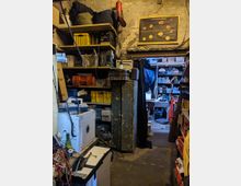 Ein kleiner, vollgestellter Werkstattraum mit Regalen, auf denen verschiedene Werkzeuge, Kisten und Gegenstände gelagert sind. Im Hintergrund befindet sich ein Durchgang mit einem Vorhang, der zu einem weiteren, ähnlich eingerichteten Raum führt. Wand- und Bodenflächen wirken abgenutzt.