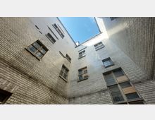 Blick in einen engen Innenhof eines Gebäudes mit Wänden aus weißen Klinkersteinen und mehreren rechteckigen Fenstern. Der Himmel ist teilweise sichtbar, und die Fenster zeigen teils beschädigte oder mit Holzplatten abgedeckte Scheiben.