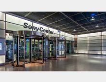 Eingangsbereich des Sony Centers mit zwei gläsernen Drehtüren, umgeben von einer modernen, beleuchteten Fassade aus Milchglas und Metall. Im Inneren sind Rolltreppen und ein Schild mit der Aufschrift "food & shops" zu sehen.