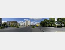 Straßenkreuzung in einem urbanen Gebiet mit mehrstöckigen Wohn- und Geschäftsgebäuden auf der linken Seite, einem modernen weißen Hochhaus im Hintergrund und großen Bäumen auf der rechten Seite. Der Himmel ist blau mit vereinzelten weißen Wolken, und es gibt wenig Verkehr auf der Straße.