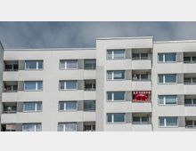 Mehrstöckiges Wohngebäude mit einer modernen grauen Fassade und rechteckigen Fenstern. Ein Balkon ist mit einem roten Sonnenschirm und bunten Lichterketten dekoriert, während eine Person auf einem anderen Balkon steht.