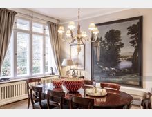 Esszimmer mit einem großen Holztisch und passenden Stühlen, einem Kronleuchter mit Lampenschirmen und einem großen Wandgemälde, das eine ländliche Szene zeigt. Links befindet sich ein großes Fenster mit Vorhängen, das Tageslicht in den Raum lässt.