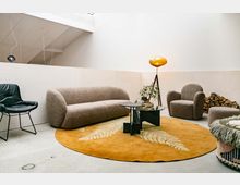 Helles Wohnzimmer mit modernen Möbeln, darunter ein beige-farbener Sessel und ein Sofa aus Bouclé-Stoff, ein schwarzer Ledersessel und ein runder, orangefarbener Teppich mit Farnmuster. Ein Couchtisch mit Glasplatte steht in der Mitte, daneben eine gelbliche Stehleuchte auf einem Stativ, im Hintergrund gestapeltes Brennholz.