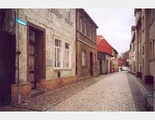 Eine schmale gepflasterte Straße in einer Altstadt, gesäumt von alten Häusern mit verputzten Fassaden und roten Ziegeldächern. Im Hintergrund sind weitere Gebäude und Bäume zu sehen.