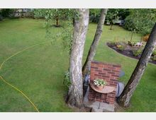 Grüne Gartenlandschaft mit Rasenfläche, mehreren Bäumen und einer kleinen gemauerten Gartenfeuerstelle aus roten Ziegelsteinen. Im Hintergrund sind Beete mit Blumen und ein Zaun zu sehen.
