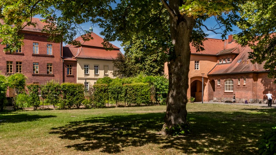 Innenhof mit Rasenfläche und großen, schattenspendenden Bäumen, umgeben von Backstein- und Putzgebäuden mit roten Ziegeldächern. Im Hintergrund sind Fensterreihen, ein Torbogen und Hecken entlang eines Weges zu sehen.
