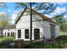 Ein weiß gestrichenes, ländliches Gebäude mit Satteldach und großen Fenstern, umgeben von Bäumen und einem Garten mit Blumen. Im Vordergrund ein Holzzaun und ein kleines Schild mit der Aufschrift "Um die Ecke".