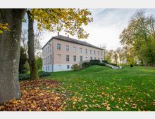 Historisches, zweistöckiges Gebäude mit rosa Fassade und weißen Fenstern, umgeben von einer gepflegten Grünfläche mit Laubbäumen und Herbstlaub auf dem Boden. Im Vordergrund sind ein Baumstamm und leicht hügeliger Rasen zu sehen.