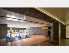 Unterirdischer U-Bahn- oder S-Bahn-Bereich mit Rolltreppen und einem Handlauf, die zu einer tieferen Ebene führen. Ein gelbes Schild mit der Aufschrift "Sony Center" und ein grünes Symbol für die S-Bahn geben Orientierung, während eine Person durch den Bereich geht.