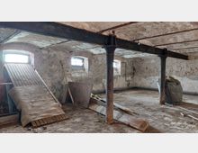 Ein leerer, verlassener Kellerraum mit bröckelnden Putzwänden, niedrigen Gewölbedecken und teilweise sichtbaren Stahlträgern. Auf dem Boden liegen Holzplatten und Schutt, daneben stehen Säulen und eine geneigte Holzrampe unter kleinen Fenstern mit Gitterstäben.