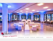 Ein moderner Veranstaltungsraum mit großen Fenstern, die den Blick auf eine städtische Umgebung ermöglichen. Der Raum ist mit weißen, minimalistischen Sitzelementen und hohen Stehtischen ausgestattet, in kühlem blauen Licht beleuchtet.