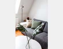 Wohnzimmer mit einer grauen Couch, dekoriert mit beigen und grünen Kissen. Im Vordergrund steht ein weißer Couchtisch mit einer Glasvase und grünen Blättern, im Hintergrund eine Stehlampe, ein Beistelltisch mit Pflanzen und eine abgeschrägte Wand mit Dachfenster.