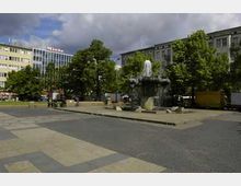 Ein öffentlicher Platz mit einem Brunnen in der Mitte, umgeben von Bäumen und mehrstöckigen Gebäuden, darunter ein modernes Gebäude mit großen Glasfenstern und dem Schriftzug „Nicole“.