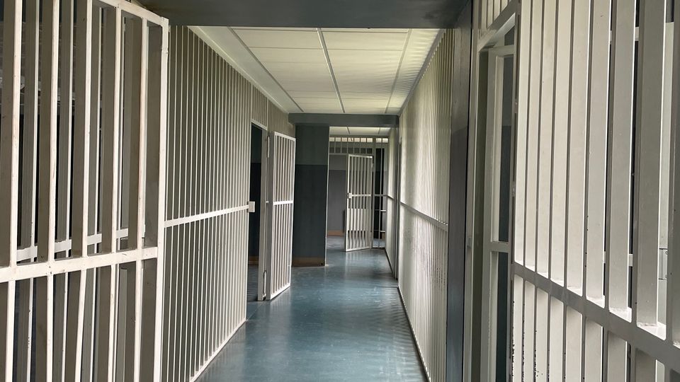 Langer Flur in einem Haft- oder Gefängnistrakt mit blauem Boden, abgehängter Decke und mehreren offenen Gittertüren entlang der Seiten. Die Sicht führt zu weiteren Gitterabschnitten und Türen am Ende des Korridors.