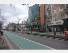 Straßenszene mit mehrstöckigen Gebäuden, darunter ein modernes Gebäude mit Glasfassade und angrenzenden historischen Häusern mit verzierten Fassaden. Vor den Geschäften verlaufen Fahrradwege und Gehwege, die von Bäumen gesäumt sind.