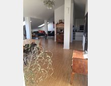 Helles Wohnzimmer mit offenem Grundriss und Dachschrägen, ausgestattet mit Holzboden, einer Sitzecke mit schwarzem Sofa, einem Esstisch und einer kleinen Küche im Hintergrund. Ein antiker Holzsekretär steht an einer weißen Wand, und eine moderne Deckenleuchte ist sichtbar.