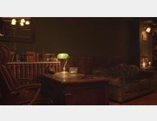 Ein gemütliches Büro mit dunklen Holzmöbeln, einschließlich eines Lederschreibtischstuhls, eines Holzschreibtisches mit einer grünen Schreibtischlampe und einer Ledercouch. Im Hintergrund befinden sich ein Regal mit Büchern und Dekorationsgegenständen sowie eine gedämpfte Beleuchtung.