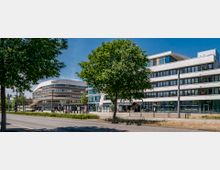 Moderne Büro- und Geschäftsgebäude mit großen Glasfronten entlang einer Straße; im Vordergrund befinden sich Bäume, ein Gehweg und eine Haltestelle mit Wartehäuschen. Im Hintergrund ist ein abgerundetes Gebäude zu sehen, das durch seine architektonische Form auffällt.