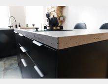 Moderne Küche mit einer Arbeitsplatte aus Beton und schwarzen Unterschränken mit matten Griffen. Im Hintergrund sind ein Wasserhahn, Fenster und Regale mit Dekoration zu sehen.