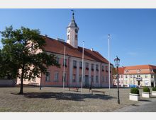 Historisches Gebäude mit rosa Fassade, rotem Ziegeldach und einem Uhrenturm auf einem gepflasterten Platz. Im Vordergrund stehen eine Laterne sowie Bänke, im Hintergrund weitere Häuser mit pastellfarbenen Fassaden.
