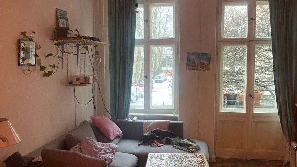 Gemütliches Wohnzimmer mit grauem Ecksofa, niedrigem Couchtisch und Holzstuhl auf Parkettboden. Große, hohe Fenster und eine Balkontür mit Vorhängen lassen Tageslicht herein; an der Wand hängen Regale mit Pflanzen und Dekoration.