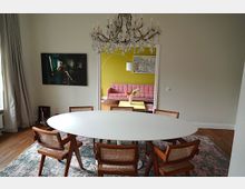 Esszimmer mit ovalem, weißem Tisch und sechs Stühlen mit Holzrahmen und geflochtener Sitzfläche, auf einem Teppich mit blau-grünem Muster. Im Hintergrund ein Durchgang zu einem Wohnzimmer mit gelber Wand, rosafarbenem Sofa und moderner Dekoration.