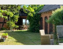 Grüne Gartenlandschaft mit einem alten Backsteingebäude und einem Fachwerkbau, teilweise von Efeu bewachsen. Im Vordergrund stehen Gartenstühle, während unter einem offenen Holzvordach Gartenmöbel sichtbar sind.