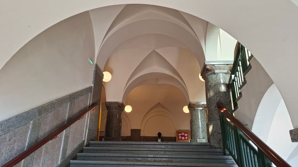 Innenansicht eines Treppenhauses mit breiter Steintreppe, dunklen Granitsäulen und einem hohen, weiß verputzten Kreuzgewölbe. Runde Wandlampen und ein grünes Geländer rahmen den Aufgang zu einem gewölbten Flur.